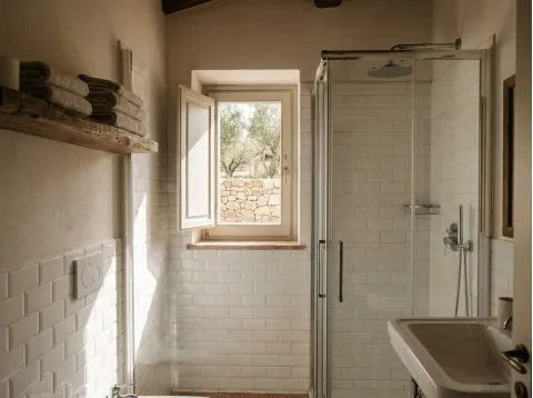 Bagno della Camera Ginepro con piastrelle bianche, box doccia in vetro e finestra sugli ulivi