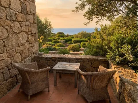Terrazzino con due poltrone in vimini e un tavolino di legno, con il giardino e il mare al tramonto