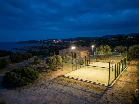 Campo da padel illuminato di sera accanto a un edificio in pietra, con le luci di un paese lungo la costa