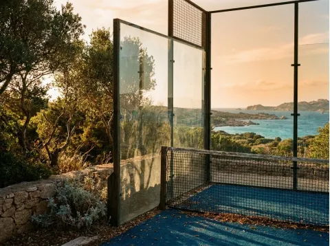 Angolo del campo da padel con parete in vetro, rete e fondo blu, e la costa al tramonto