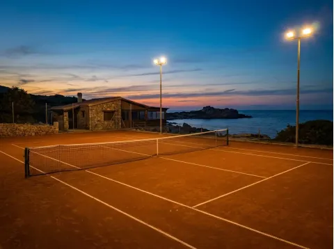 Campo da tennis illuminato dai fari al tramonto, con un edificio in pietra e il mare sullo sfondo